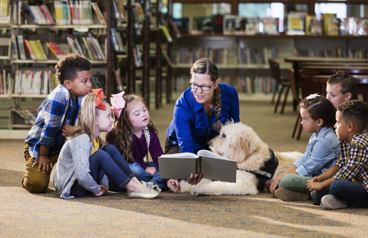A multi-ethnic group of six boys and girls sitting on the floor of a library, reading with a therapy dog, a goldendoodle. The dog handler is a mature woman in her 50s who is smiling holding a book and reading aloud. The girl 3rd from the left has down syndrome.